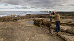 Photographer with telephoto lens on rocky coastal beach overlooking water and distant shoreline.
