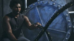 Man in dark tank top sits against large blue industrial gear wheel with metal studs and rivets.