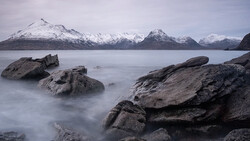 Snow-capped mountains across a calm fjord with foreground rocks smoothed by long-exposure water effect.