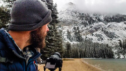 Bearded photographer in winter beanie holding camera equipment overlooking snow-covered alpine lake and mountains.