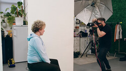 Photographer adjusting camera settings while shooting a portrait of a person seated against a white wall in a studio setting.