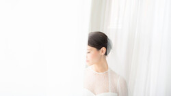 Woman in profile wearing a light knit sweater, standing near sheer white curtains with soft natural light.