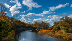 River flowing through autumn landscape with golden trees and blue sky with white clouds.