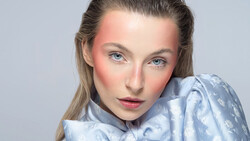 Portrait of a woman with blue eyes and pink-toned cheeks against a neutral gray background.