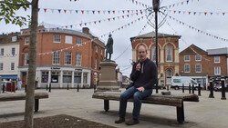 Person sitting on a wooden bench in a town square surrounded by Victorian-era brick buildings and festive bunting.