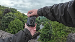 Hands holding a thermal imaging camera pointed at a lush green valley landscape with a river visible below.
