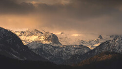 Snow-capped mountain peaks illuminated by golden light breaking through storm clouds above a forested valley.