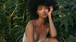 Woman with voluminous natural hair wearing a cream-colored top, posed among lush green foliage.