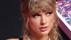 Close-up portrait of a woman with blonde hair and bangs, wearing dramatic makeup and gold chain necklace.