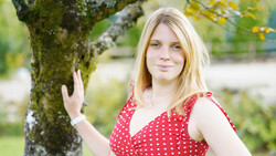Young woman in red polka dot dress posing with hand on moss-covered tree trunk in garden setting.