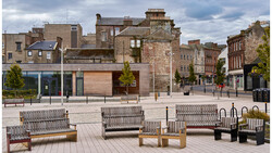 Contemporary public plaza with metal benches and wooden seating arranged on light-colored pavement, facing historic stone buildings.