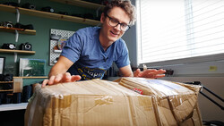 Photographer unwrapping a large brown cardboard package in a bright studio workspace.