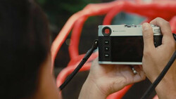 Photographer holding a rangefinder camera with a wrist strap while wearing a red jacket.