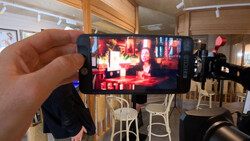 Hand holding smartphone displaying video content in a studio setting with red lighting and seated figures.
