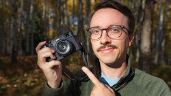 Male photographer holding a rangefinder camera while standing in an autumn forest setting.