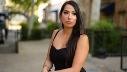 Woman in black sleeveless dress posing outdoors on urban street with blurred background.