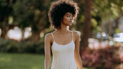 Woman in white camisole dress posing outdoors among trees and grass.