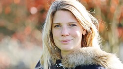 Woman with blonde hair wearing dark jacket with fur collar, photographed outdoors during golden hour.