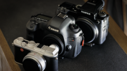Three digital cameras arranged on a dark surface: a Canon EOS 5D Mark IV, a Fujifilm X-T4, and a compact mirrorless camera in silver.