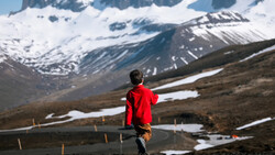 Runner in red jacket jogging on mountain road with snow-capped peaks and patches of snow in background.