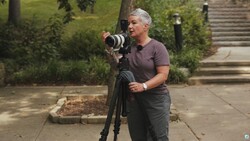 Photographer holding a telephoto lens on a tripod while composing a shot outdoors.