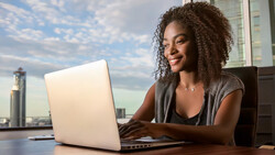 Woman working at laptop at desk with city skyline visible through window behind her.