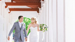 Bride and groom walking together through a white columned corridor holding hands.