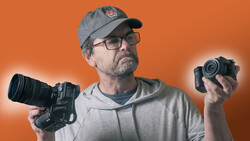 Videographer holding cinema camera in left hand and compact camera in right hand against orange background.