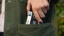 Hand holding a compact rangefinder camera with white body and black lens protruding from olive cargo pants pocket.