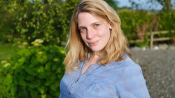 Woman with blonde hair wearing a light blue button-up shirt, photographed outdoors with green foliage in the background.