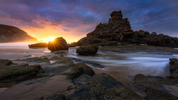 Rocky coastline at sunset with golden light between layered stone formations and smooth water in foreground.
