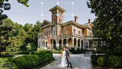 Couple in wedding attire posing in front of a brick Victorian mansion with a tower, surrounded by landscaping.