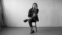 Person sitting on a wooden chair in a minimalist studio setting, resting chin on hand in a contemplative pose.