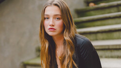 Young woman with long reddish-brown hair wearing black jacket, posed near moss-covered concrete steps.