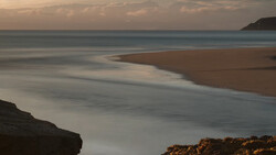Serene coastal landscape at dusk with smooth water, sandy beach, and rocky outcrops.