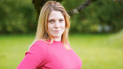 Woman in bright pink ribbed sweater poses outdoors against blurred green background.
