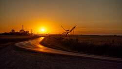 Rural road curving toward a bright sunrise, with an irrigation pivot sprinkler system silhouetted in golden light.
