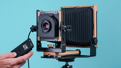 Large format camera with wooden frame and bellows, held alongside a cable release against a blue background.