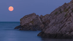 Full moon rising above rocky coastal cliffs at dusk with calm blue water below.