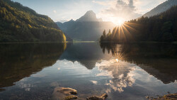 Mountain lake at sunrise with forested slopes and perfect mirror reflection in still water.