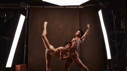Two dancers in a studio performing a contemporary lift against a brown backdrop with professional lighting.