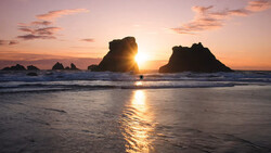 Sun setting between two large sea stacks silhouetted against golden sky and ocean.