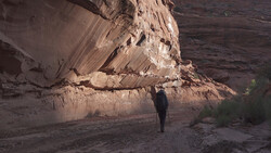 Photographer standing beneath towering sandstone cliff face in desert canyon.