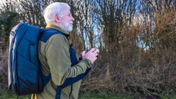 Older man with white hair wearing a blue backpack and olive shirt, holding a camera while looking into the distance amid bare trees.