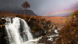 Waterfall cascading over rocky terrain with rainbow arcing across stormy sky and mountains.
