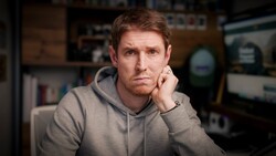 Man in gray hoodie resting his head on his hand, looking thoughtfully at camera in a studio setting.