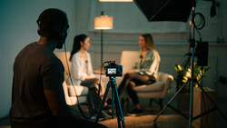Camera on tripod in foreground during a studio interview setup with two people seated in background.