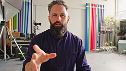 Bearded man in dark work shirt gesturing while speaking in a studio with colorful background rolls and lighting equipment.