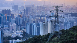 High-voltage transmission tower overlooking a sprawling cityscape at dusk with mountains in the distance.
