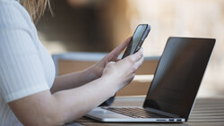 Person holding a smartphone while working at a laptop outdoors in natural daylight.